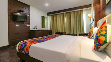 A hotel room with a bed, a TV mounted on the wall, a desk with a lamp, and a window with curtains.
