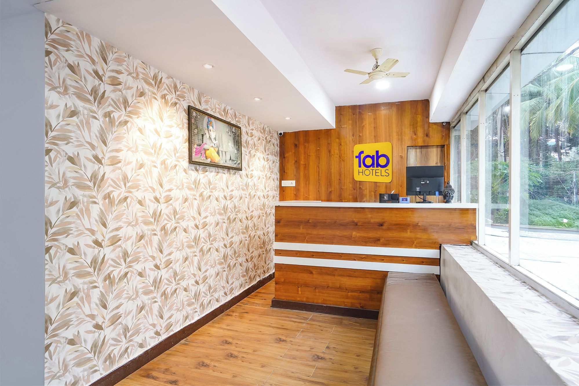 A hotel reception area with a wooden reception desk, a framed artwork on the wall, and a large window offering a view of greenery.