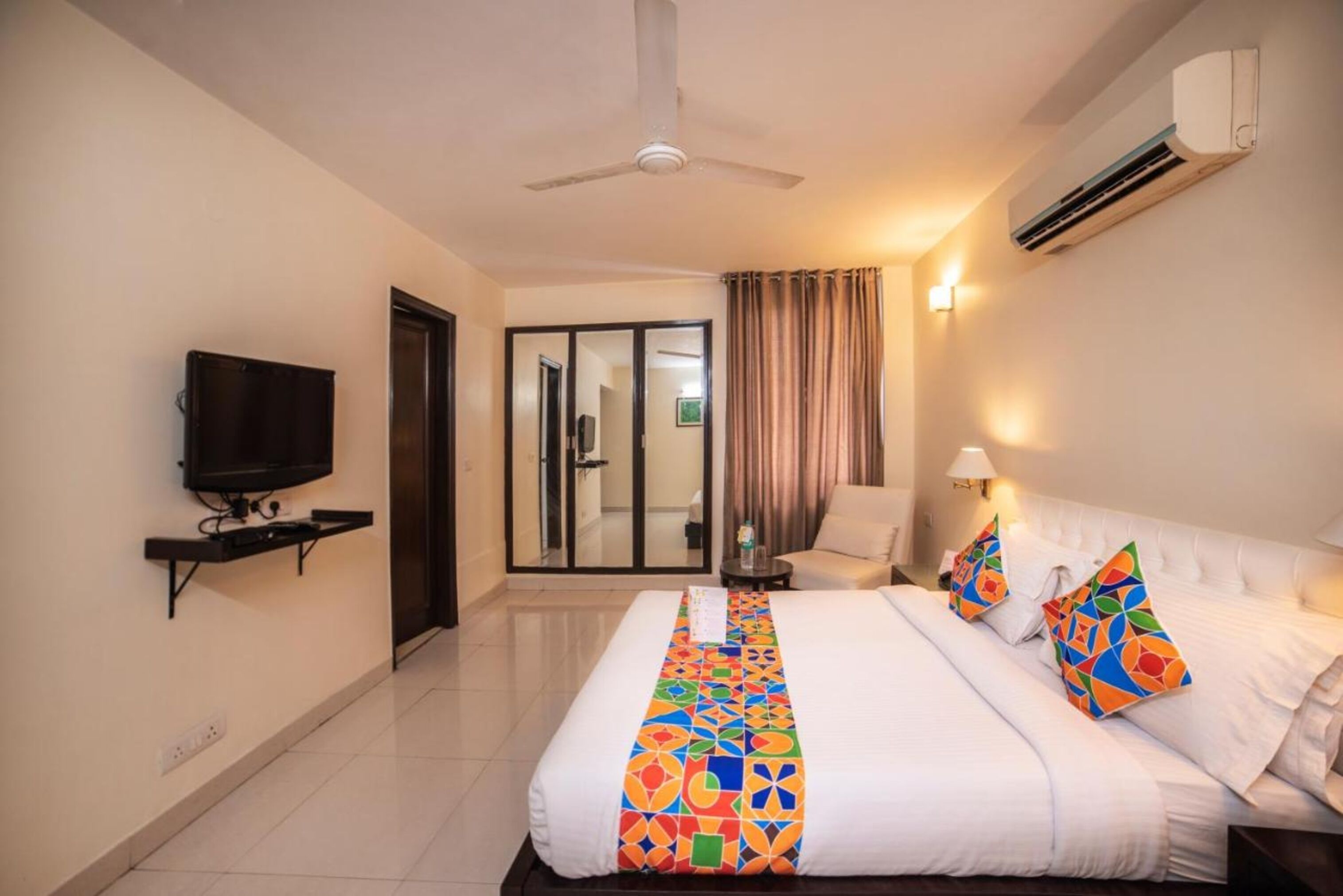 A hotel room with a bed, a TV, a ceiling fan, an air conditioner, a chair, and a sliding glass door leading to a bathroom.