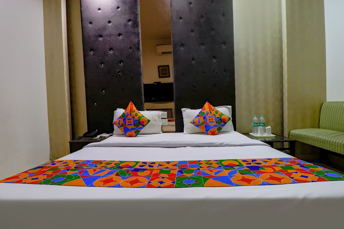A hotel room with a large bed, two pillows with colorful geometric patterns, a bedside table with water bottles, and a green chair.