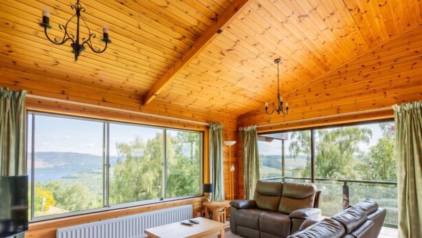 Een gezellig interieur van een blokhut met houten balkenplafonds, een leren bank, een televisie en grote ramen met uitzicht op het landschap.