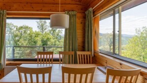 Interieur van een houten hutje met een eettafel en stoelen, grote ramen met uitzicht op bomen en heuvels.