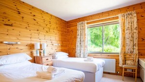 Een slaapkamer in een houten chalet met twee bedden, een nachtkastje, een stoel en een raam met gordijnen.