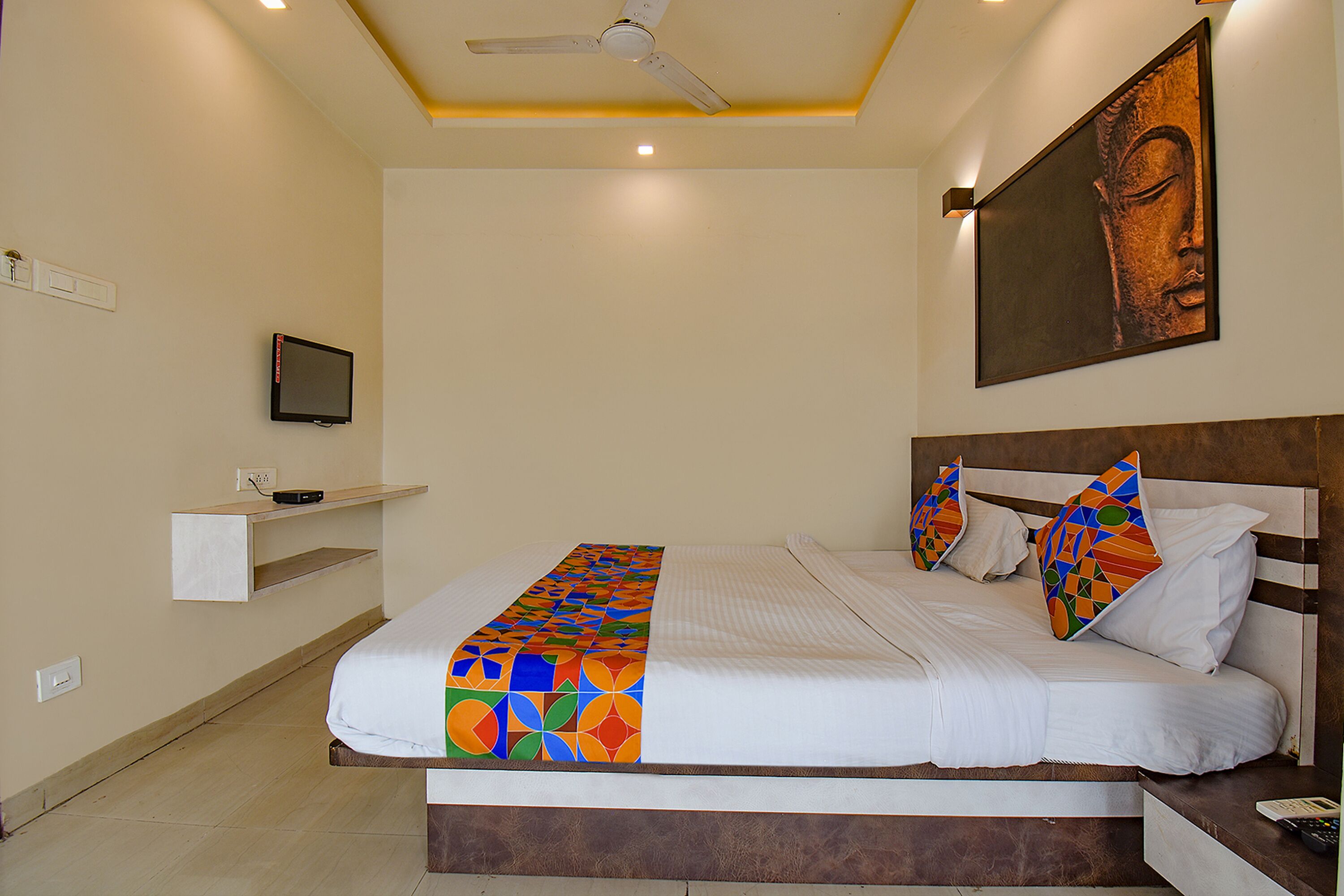 A neatly made bed with a colorful blanket, a mounted television, and a Buddha statue on the wall.