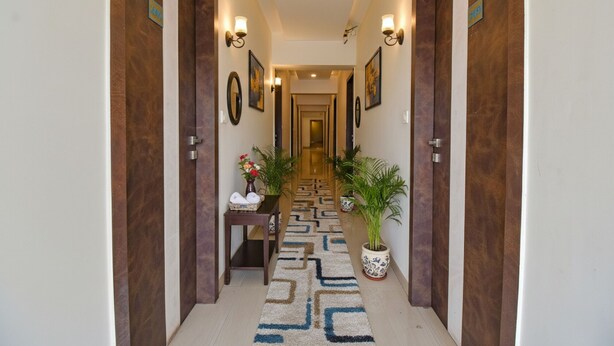 A hotel hallway with wooden doors, wall-mounted light fixtures, framed artwork, a patterned carpet, and potted plants. - FabHotel Royce Studio (Pune)