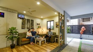 A modern living room with a wooden floor, a ceiling fan, and a television on the wall. There are three people seated on a sofa, and a small table with a magazine and a cup. - FabHotel Royce Studio (Pune)