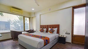 A hotel room with a large bed, a desk, a chair, a window with a view of trees, and a door with a frosted glass panel. - Fabhotel Sr Wakad (Paud)