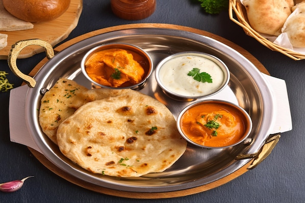 Un plato de acero inoxidable con pan naan, dos tipos de curry y un pequeño cuenco de yogur.
