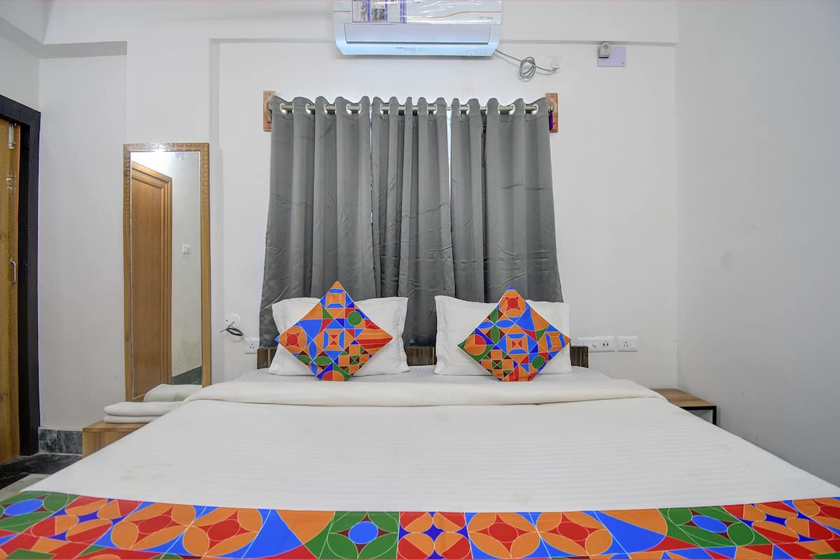 Une chambre d’hôtel avec un lit, deux oreillers, une fenêtre avec des rideaux gris, un climatiseur fixé au mur et une porte en bois.
