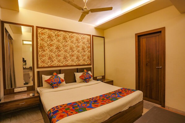 A hotel room with a bed, a nightstand, a mirror, a ceiling fan, and a wooden door. - Fabhotel Surya (Bhopal)