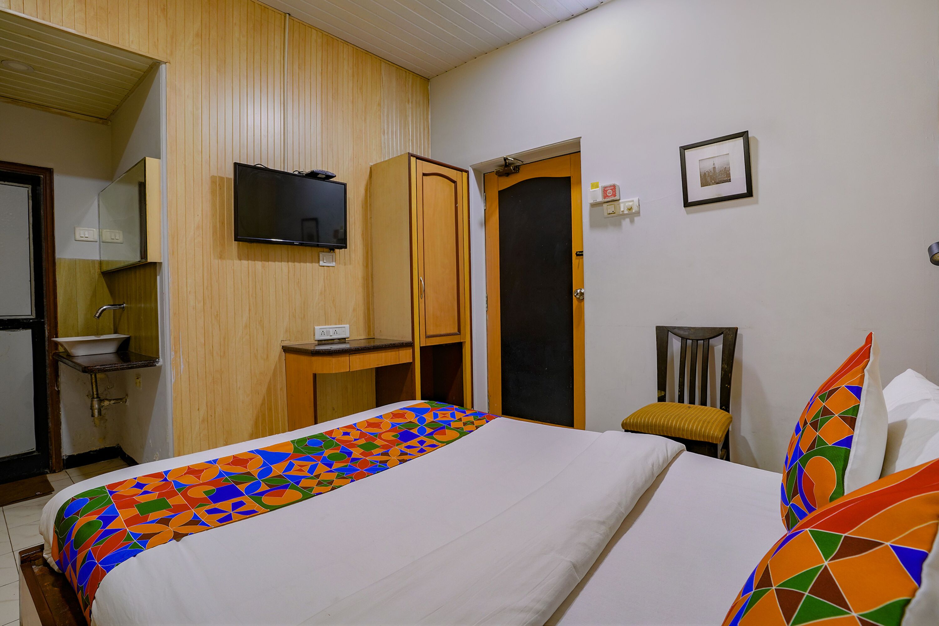 A hotel room with a bed, a wooden wardrobe, a television mounted on the wall, a small table, and a chair.