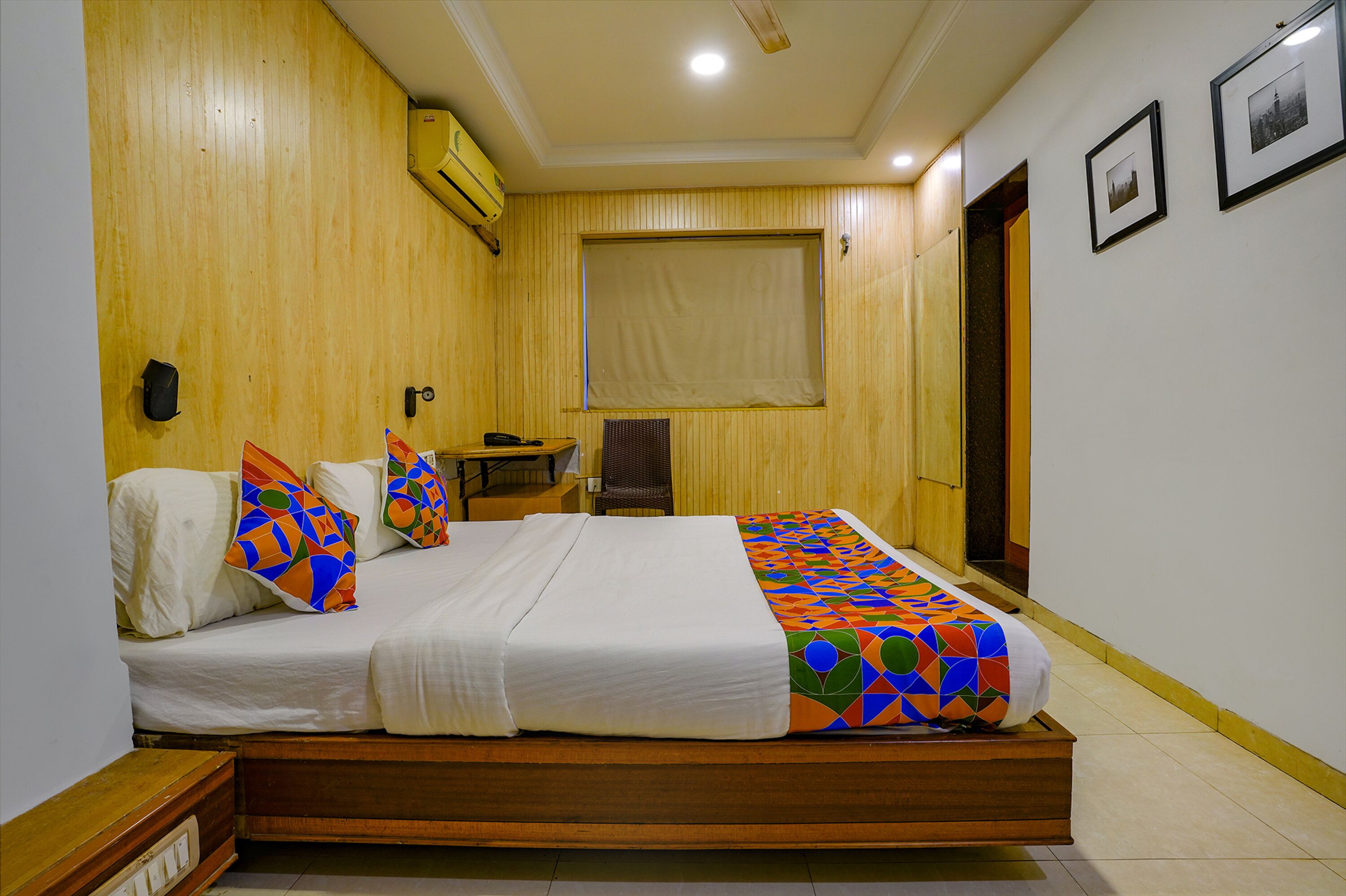 A single bed with a colorful geometric-patterned blanket, a bedside table with a lamp, and a window with a beige blind.