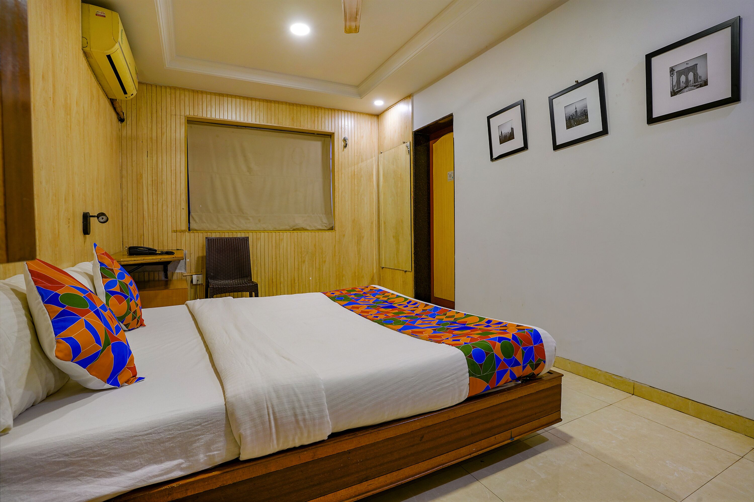 Una cama bien hecha con una manta y almohadas coloridas, un cabecero de madera, una mesita con una silla y un aire acondicionado instalado en la pared.