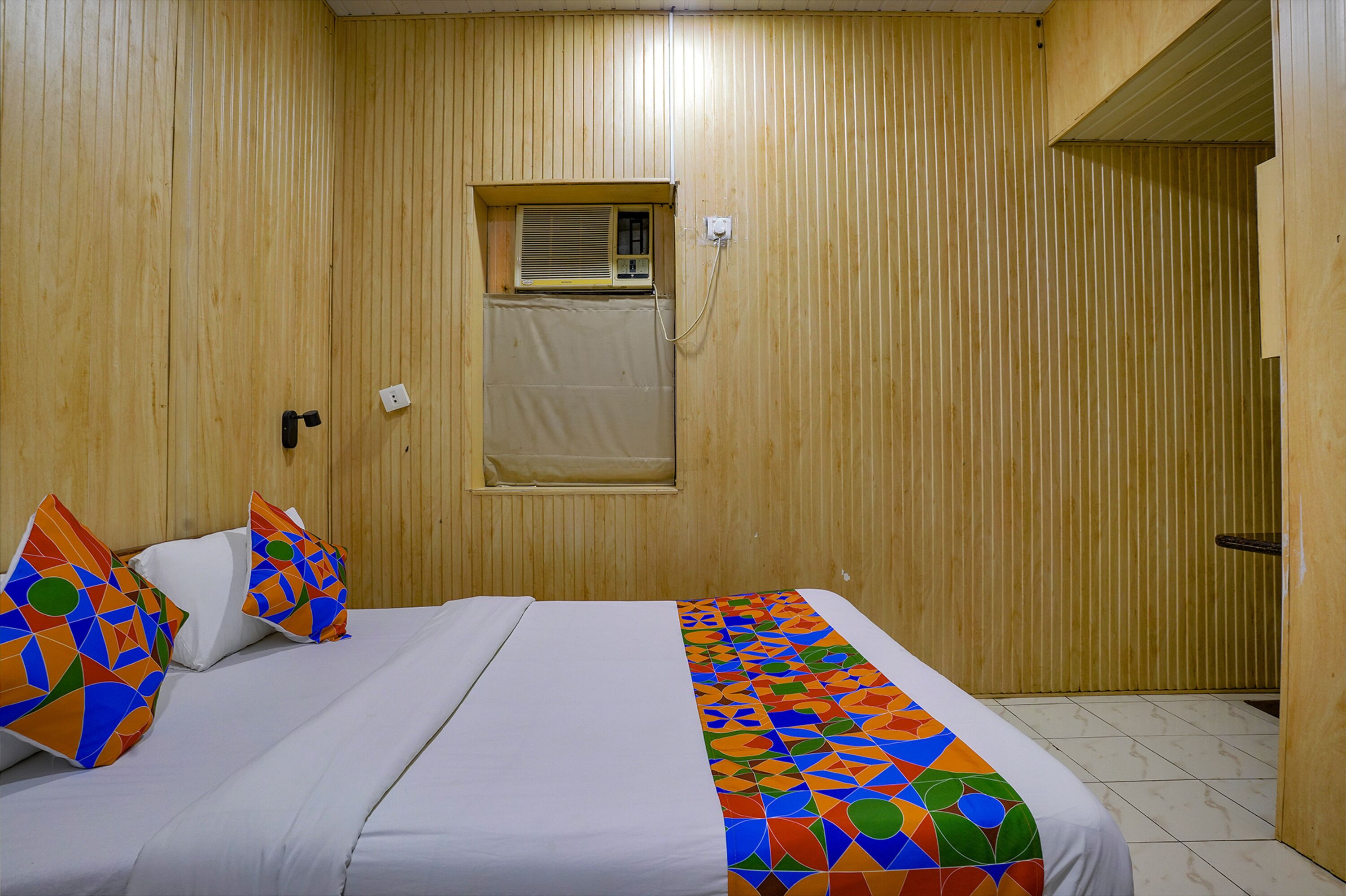 A hotel room with a wooden paneled wall, a single bed with white bedding, colorful geometric-patterned pillows, a window with blinds, and a door to the right.