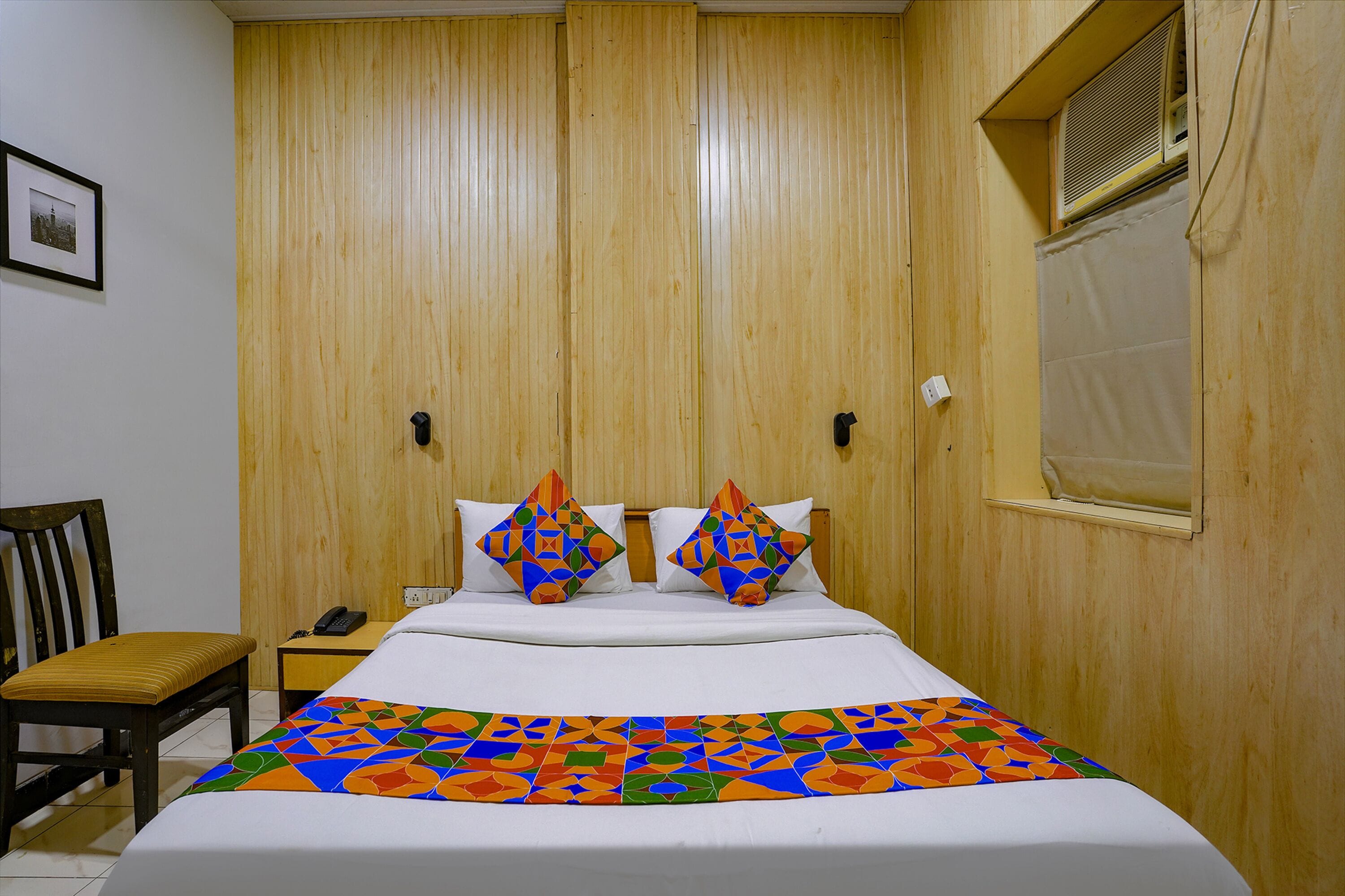 Een hotelkamer met een houten garderobe, een bed met kleurrijk beddengoed, een stoel en een nachtkastje met een telefoon.