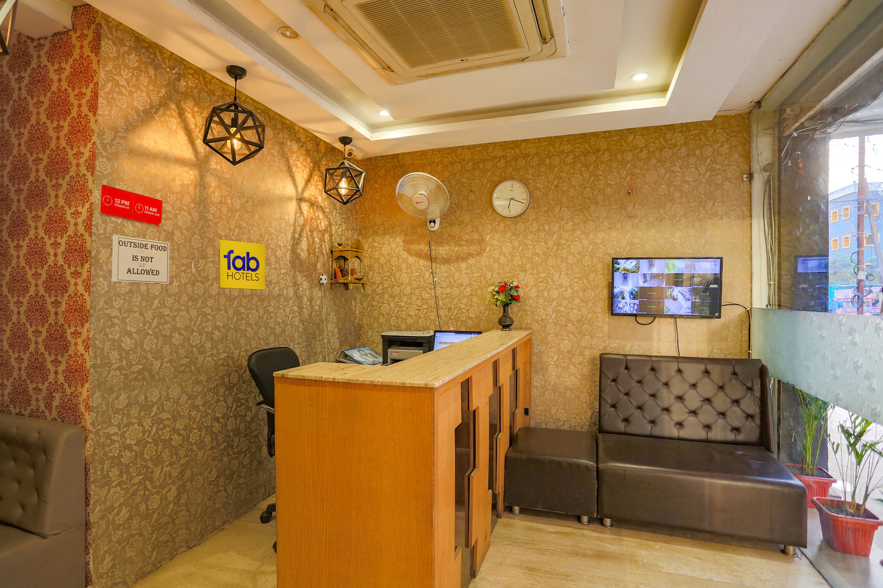 A hotel reception area with a wooden desk, a black office chair, a sofa, a clock, and a TV mounted on the wall.