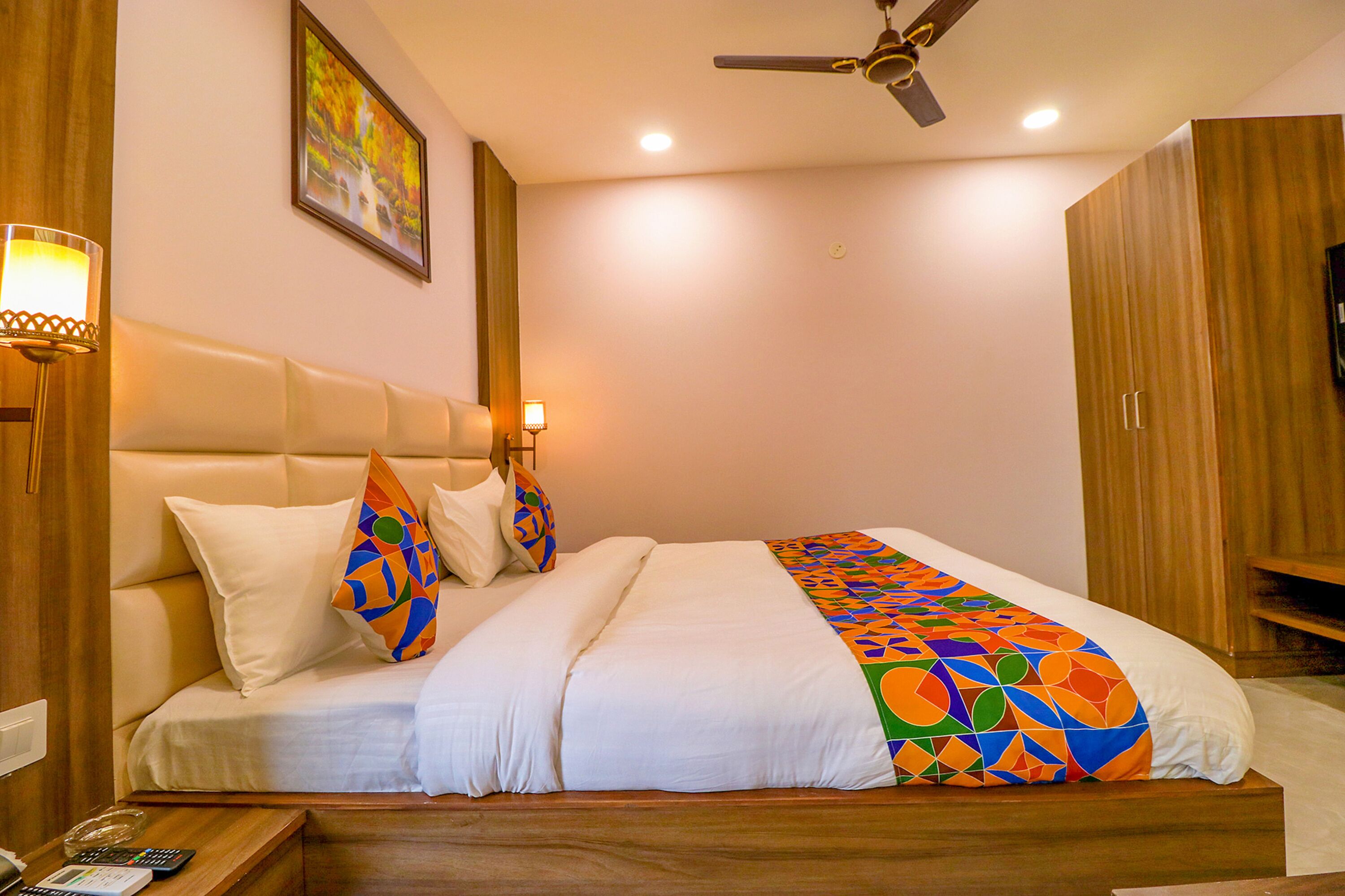 A hotel room with a bed, a nightstand, a lamp, a ceiling fan, and a painting on the wall.