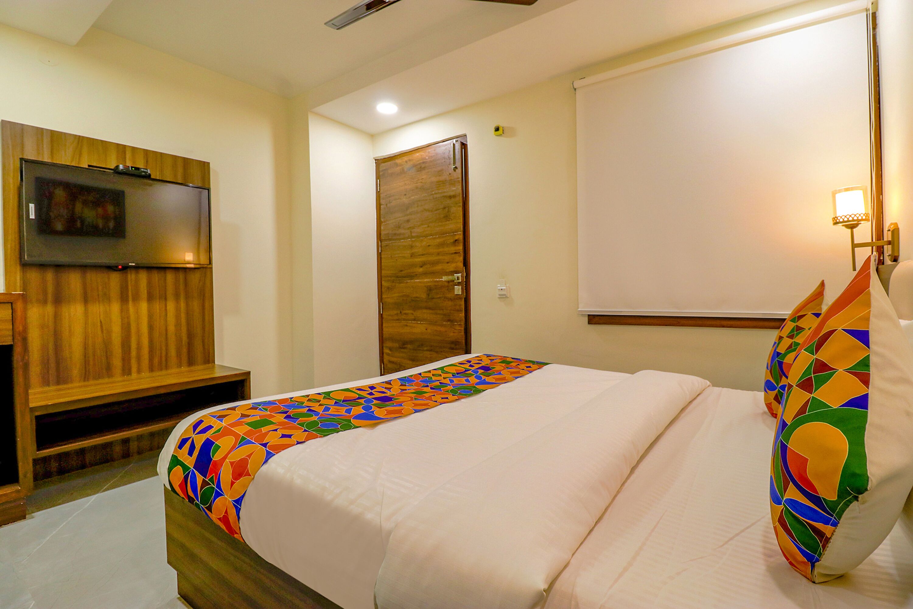 Une chambre d’hôtel avec un lit, un téléviseur fixé au mur, une porte en bois et un mur blanc agrémenté d’un luminaire.