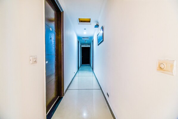 A narrow, well-lit hallway with a ceiling light, a door on the left, and a wall-mounted light fixture on the right. - Fabhotel Cameron (New Delhi)