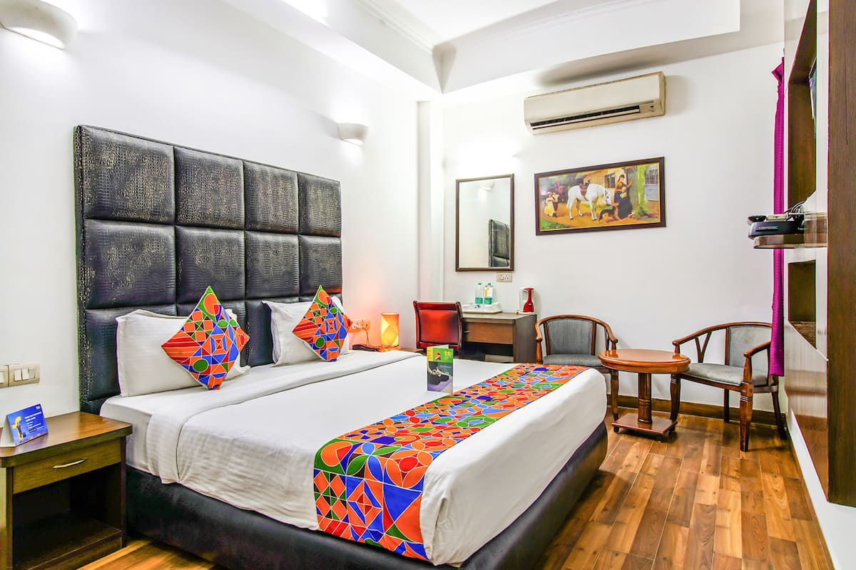 A hotel room with a large bed, a desk, two chairs, a nightstand, and an air conditioning unit.