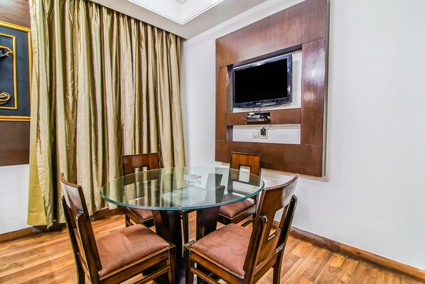 A dining area with a glass table surrounded by wooden chairs, a flat-screen TV mounted on the wall, and a large window with curtains. - Fabhotel Cameron (New Delhi)