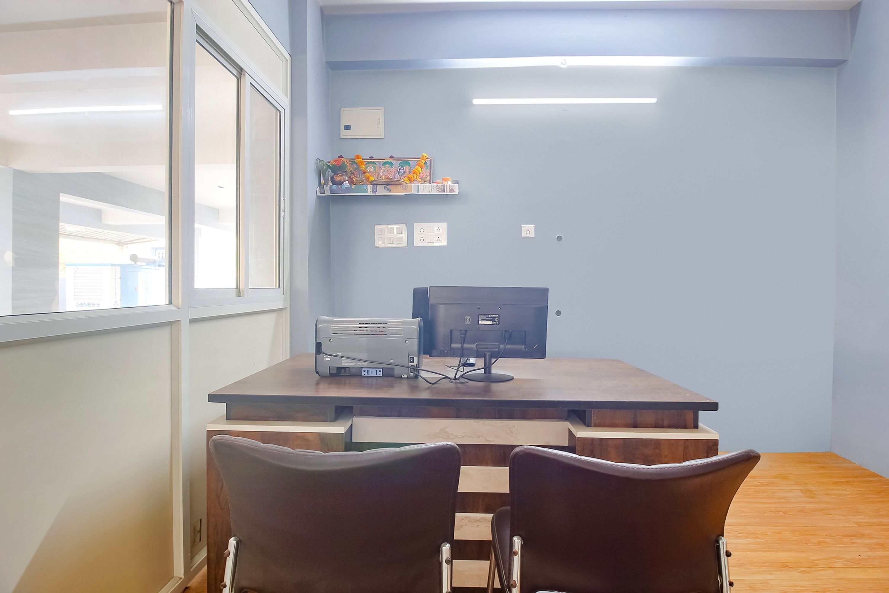 A conference room with a wooden table, two chairs, a projector, a computer, and a shelf with decorative items.