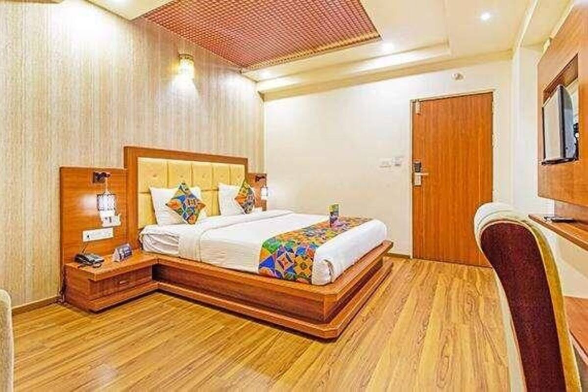 A hotel room with a wooden floor, a bed with a colorful blanket, a nightstand with a lamp, and a wooden door.