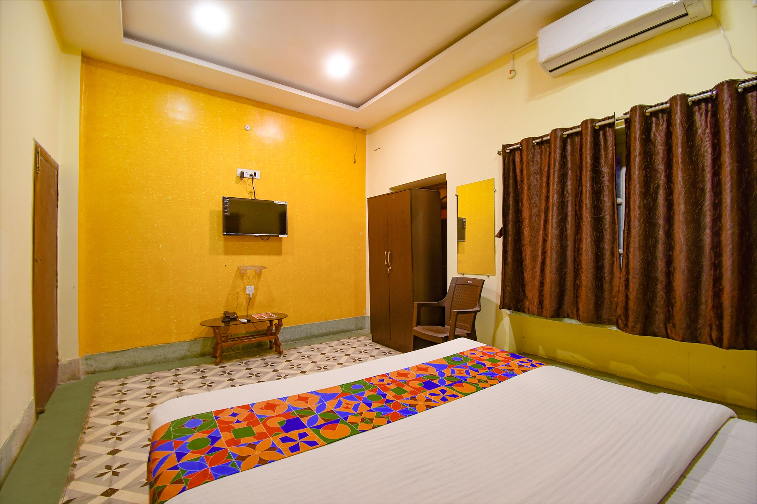 A hotel room with a bed, a television mounted on the wall, a wardrobe, and a small table with a chair.
