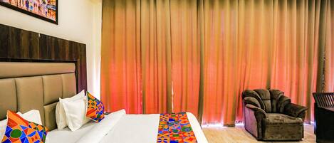 Habitación de hotel con cama, cabecera, una silla y un ventanal con cortinas.