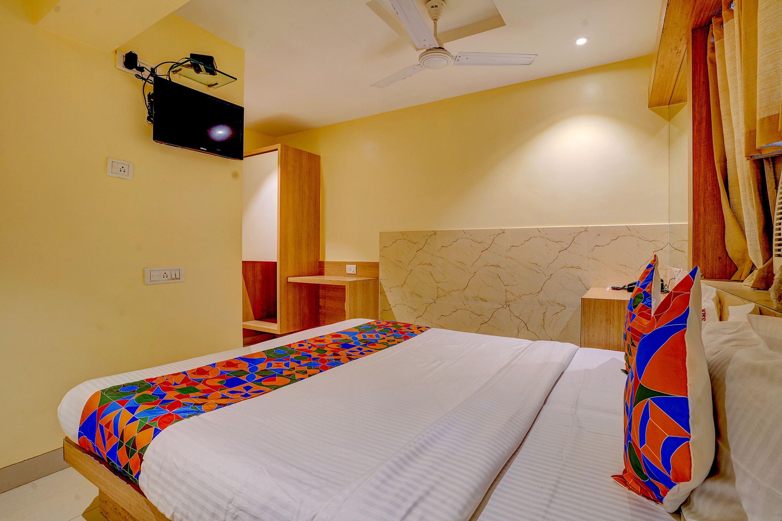 A hotel room with a bed, a ceiling fan, a TV mounted on the wall, and a wooden nightstand.