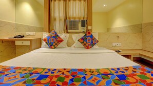 A hotel room with a large bed, two pillows with colorful geometric patterns, a bedside table with a phone, a wall-mounted air conditioner, and a mirror above the bed.