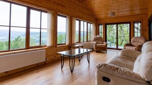 Eine geräumige Holzhütte mit großen Fenstern und reizvollem Ausblick, eine gemütliche Sitzecke mit Sofa und Sesseln sowie ein kleiner Tisch.