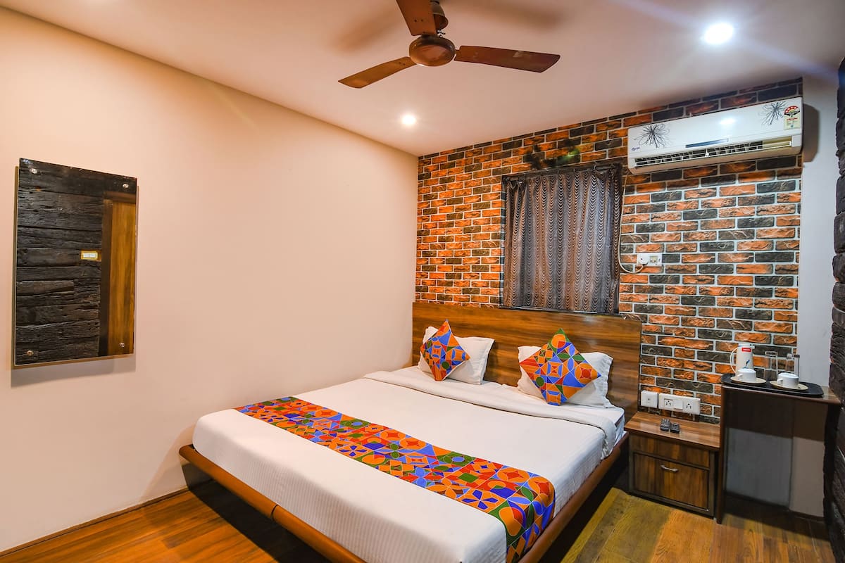 A neatly made bed with a colorful geometric-patterned bedspread, a wooden headboard, a ceiling fan, and a brick-patterned wall with an air conditioning unit.