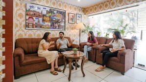 A room with patterned wallpaper, a brown sofa, a small round table, and a large window with plants outside. - Fabhotel Gargi Deluxe (Pune)