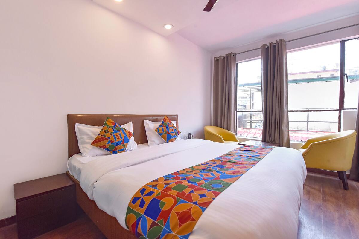 Una habitació d'hotel amb un llit gran, dos almoinos amb motius geomètrics de colors, un llençol blanc i una manta de colors. Hi ha dues cadires grogues i una finestra amb cortines.