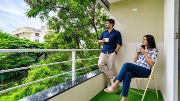A balcony with a white railing, a person standing and holding a cup, and another person seated on a chair.