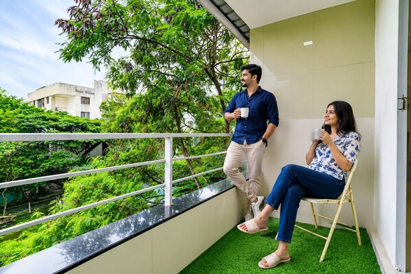 A balcony with a white railing, a person standing and holding a cup, and another person seated on a chair. - FabHotel Vardhan House I - Nr. Baner Hills (Pune)