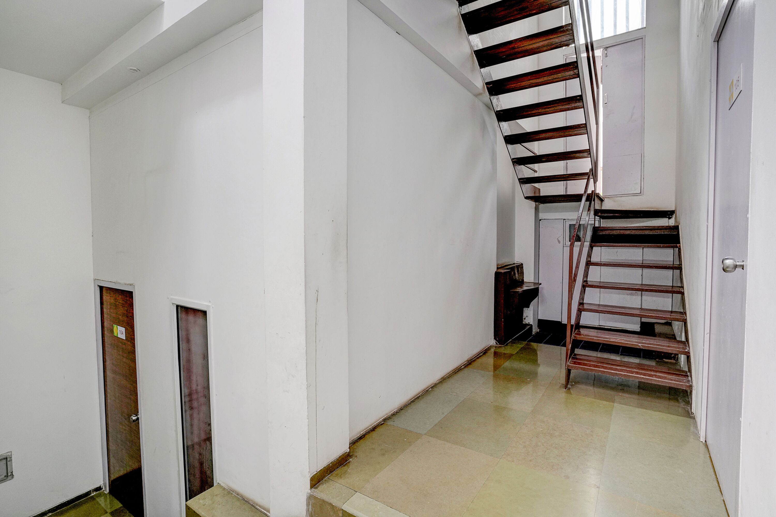A narrow hallway with a staircase on the right, a door at the end, and a trash bin against the wall.