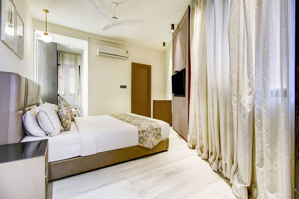 Quarto de hotel com cama, televisão, ventoinha no teto, ar condicionado e cortinas.