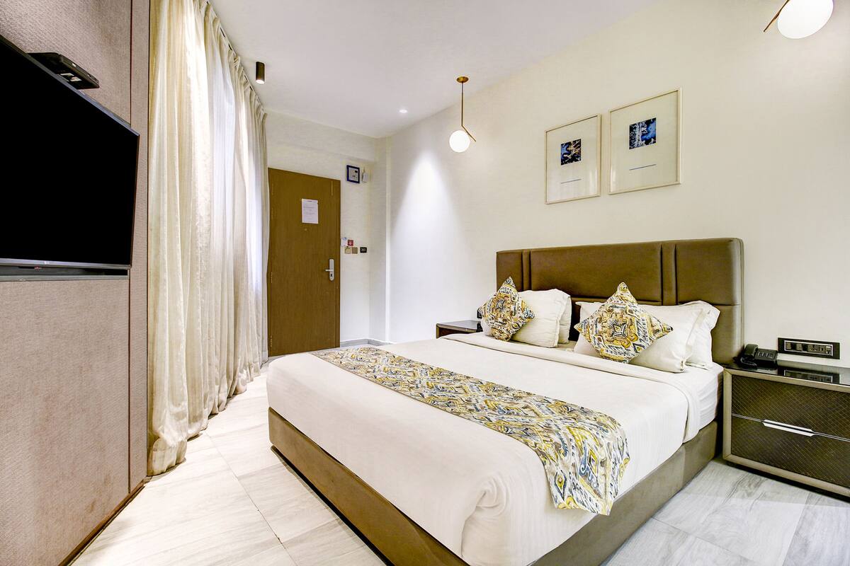 Quarto de hotel com uma cama grande, uma mesa de cabeceira com abajur, uma televisão e uma porta com placa.