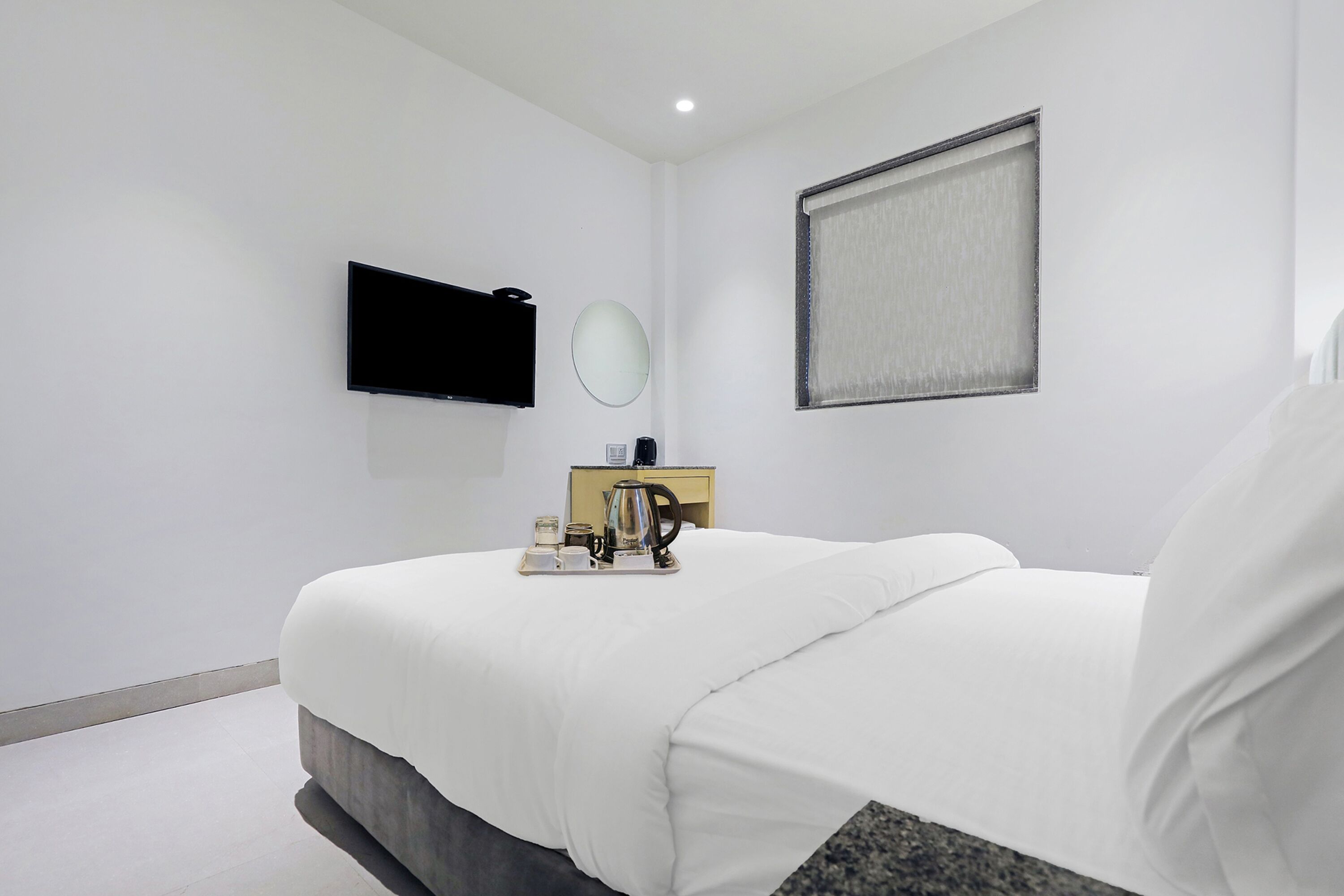A modern hotel room with a large bed, a flat-screen TV mounted on the wall, a bedside table with a kettle and cups, and a window with a blind.