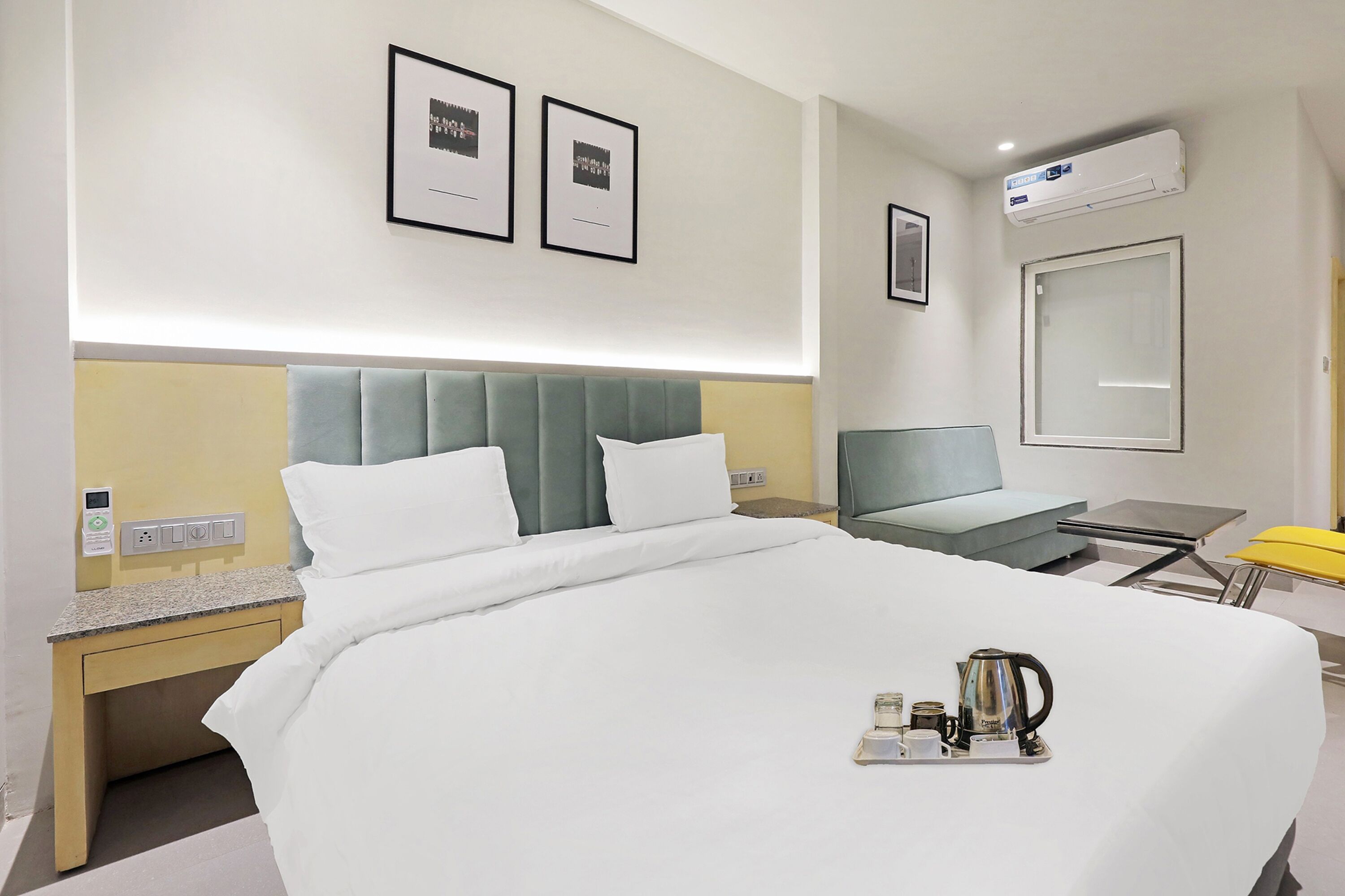 A hotel room with a bed, bedside table, and a small desk with a chair. There are framed pictures on the wall, an air conditioning unit, and a window.