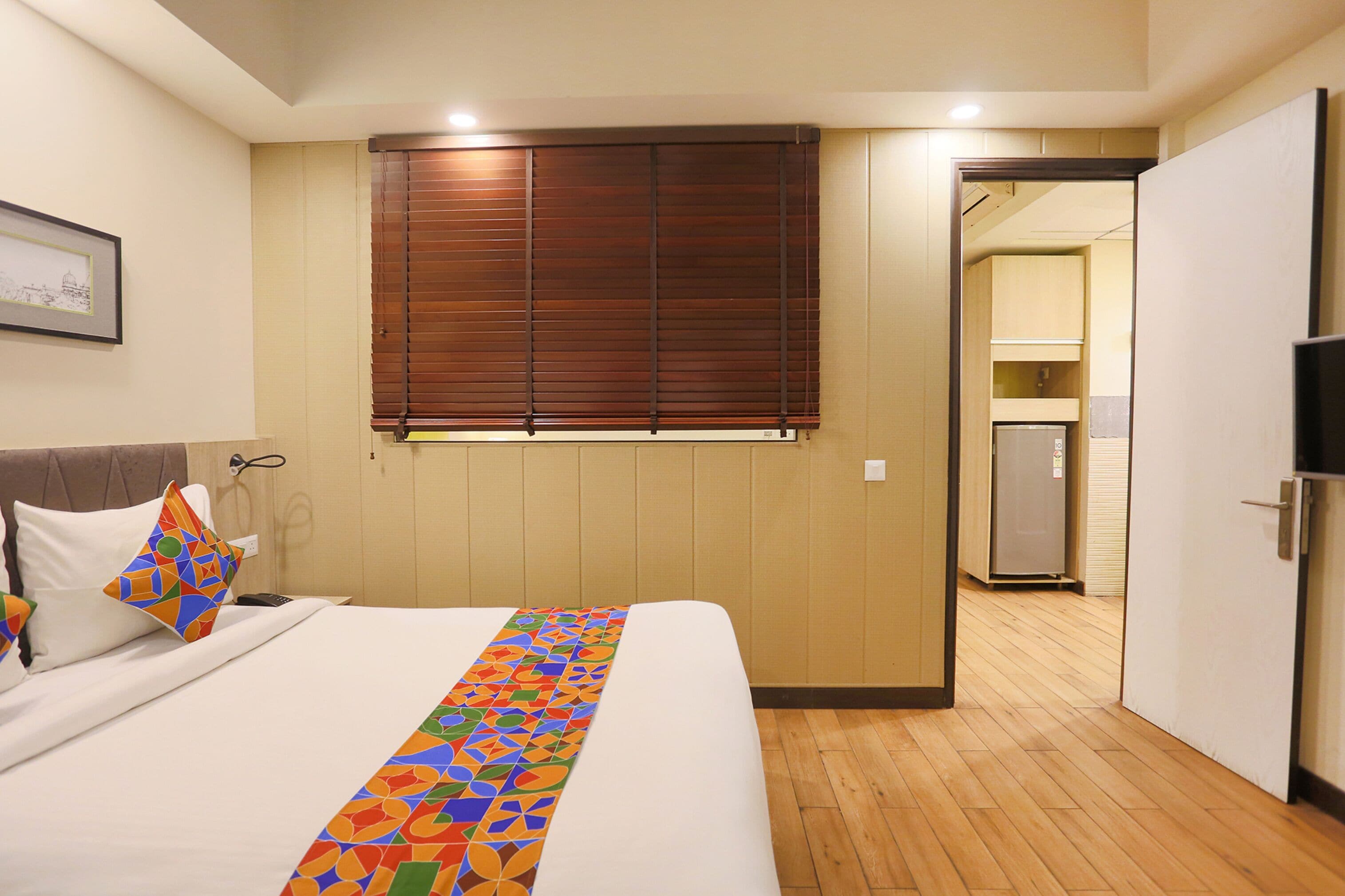 Ein Hotelzimmer mit einem Bett, einem Fenster mit Jalousien, einer Tür zu einer Küchenzeile und einem gerahmten Bild an der Wand.