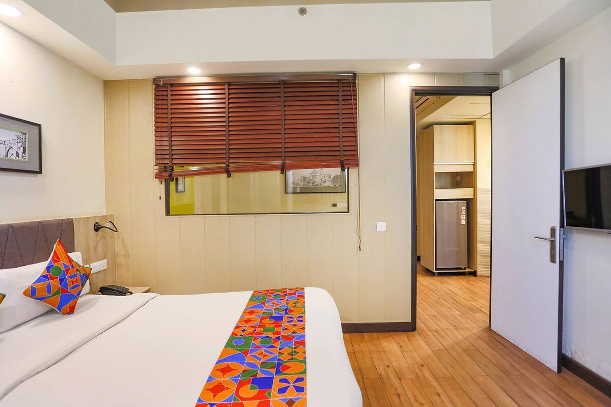 A hotel room with a bed, a window with blinds, a door leading to a kitchen, and a framed picture on the wall.