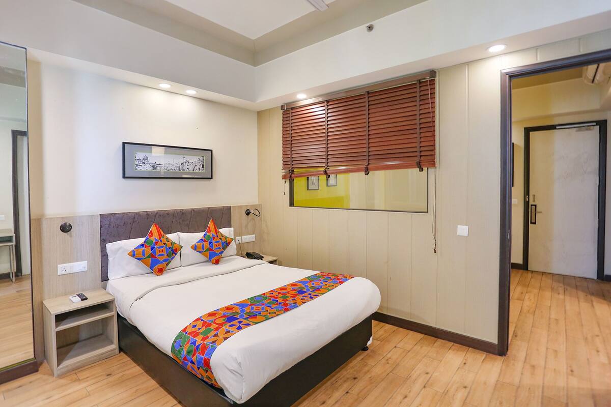A modern hotel room with a large bed, a bedside table, a nightstand, a mirror, a framed picture on the wall, and a window with blinds.