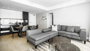 A modern living room with a grey sectional sofa, a dining area with a table and chairs, and a kitchen in the background.