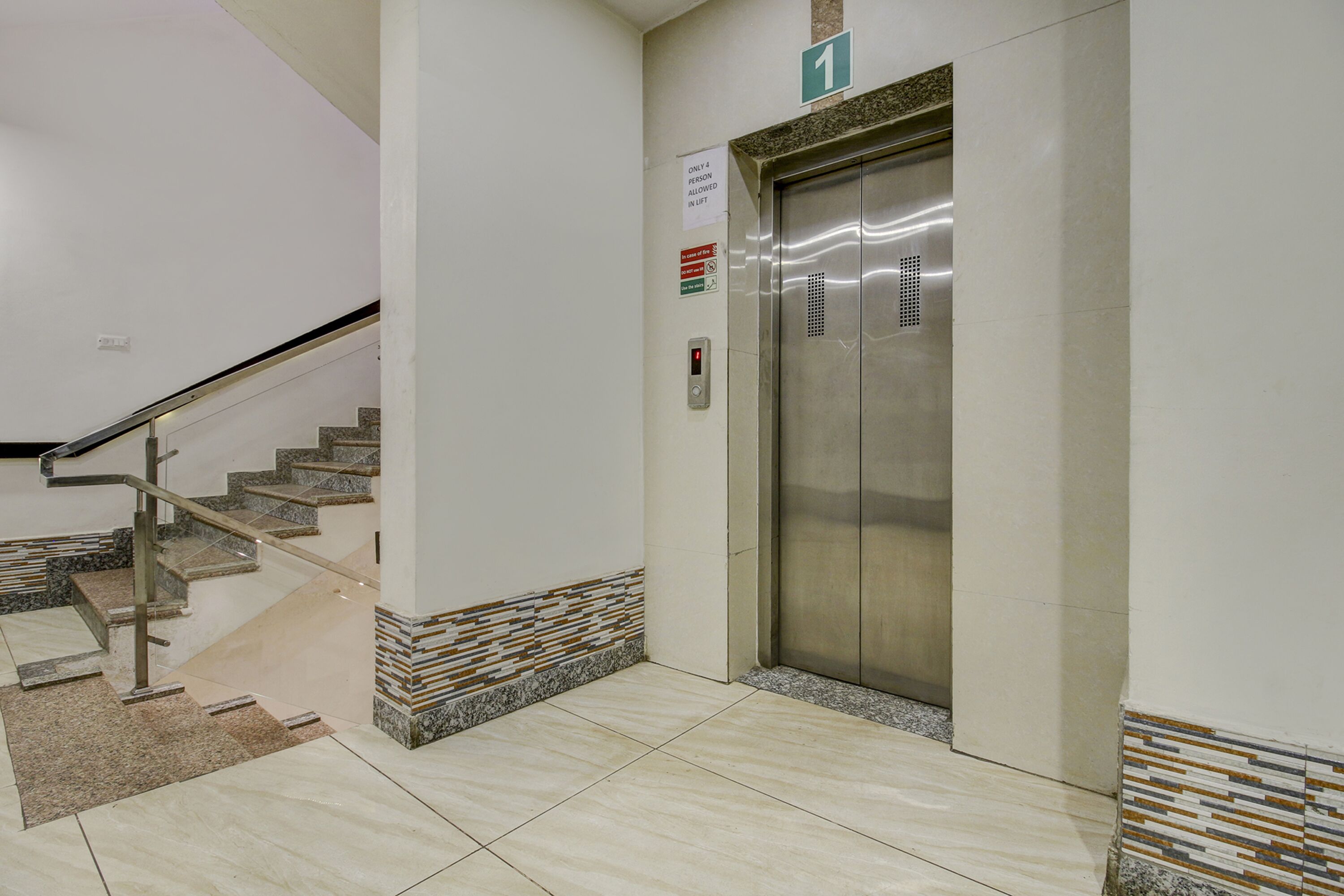 A clean, modern elevator area with a metal elevator door, a staircase with a metal handrail, and a tiled floor.