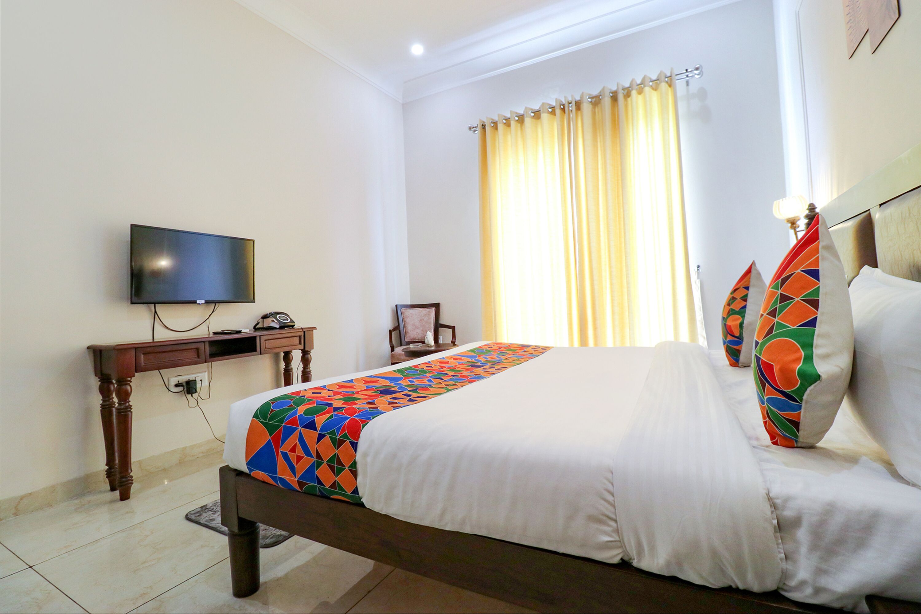 A hotel room with a bed, a desk with a television, a chair, and a window with curtains.