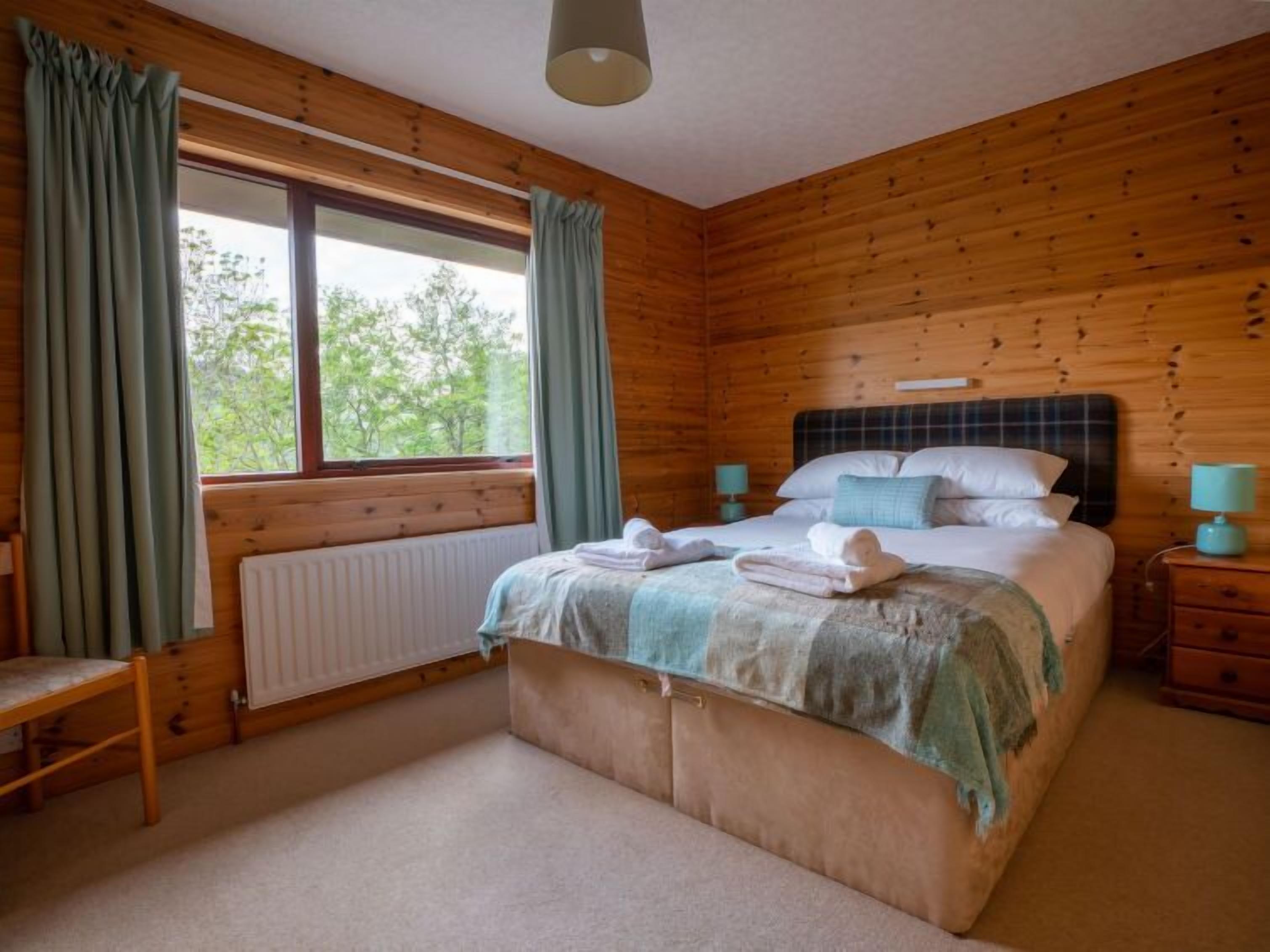 A wooden-paneled bedroom with a bed, bedside tables, a bench, and a window with curtains.