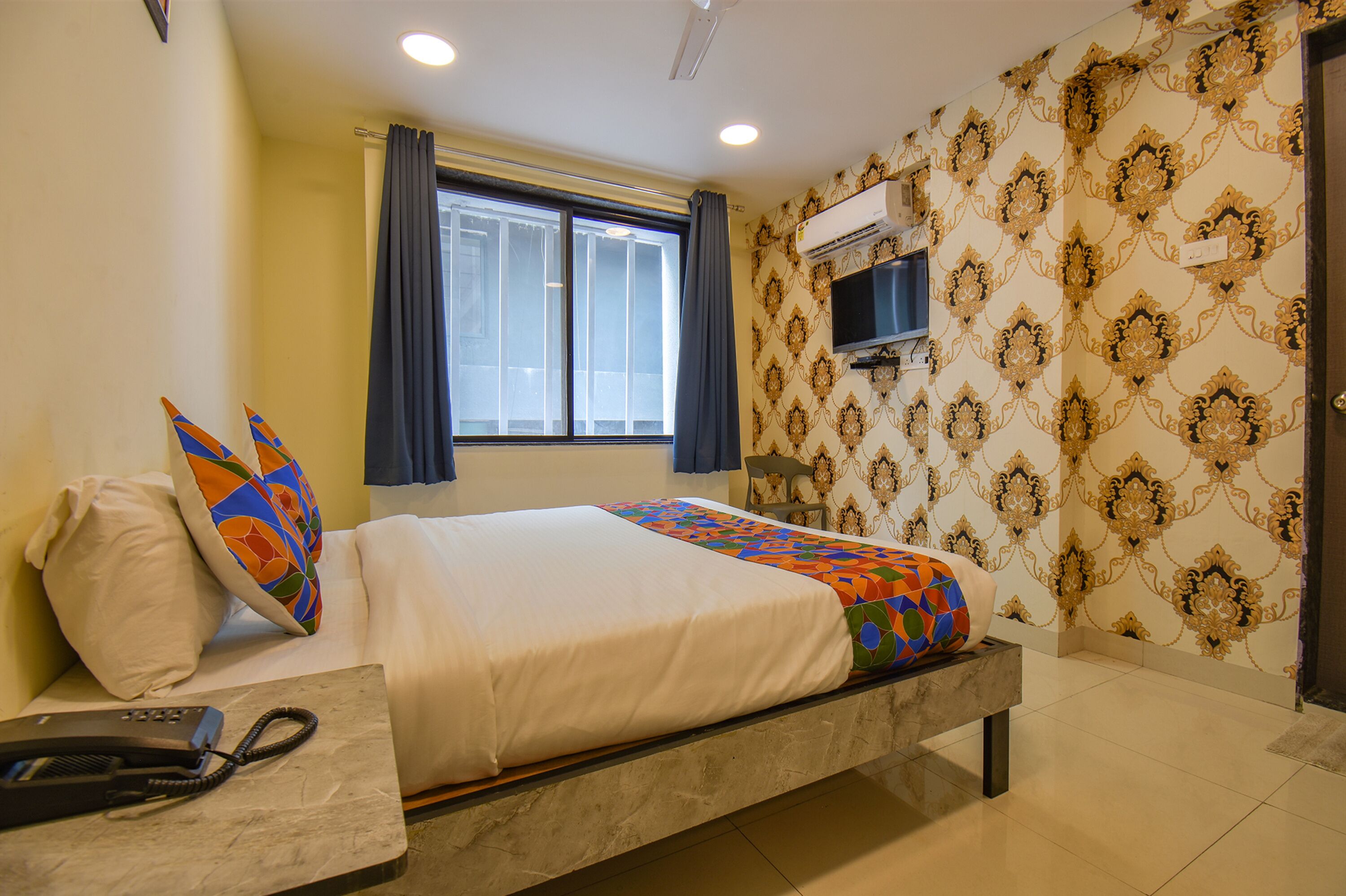 A hotel room with a bed, a telephone, a window with curtains, an air conditioning unit, and patterned wallpaper.